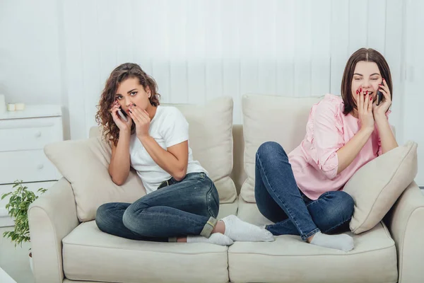Dvě úžasné dívky sedící na různých stranách pohovky a mluvící po telefonu. Jedna dívka mluví nahlas a otevřeně, druhá drží ruku u huby, mluví potichu. — Stock fotografie