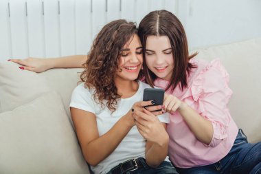 Evdeki kanepede oturan güzel genç kızlar telefondaki resimlere bakıp gülümsüyor, etkileşime geçiyorlar. Bir kız telefonu tutuyor, diğeri de ekranı işaret ediyor..