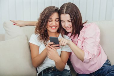 Evdeki kanepede oturan güzel genç kızlar telefondaki resimlere bakıp gülümsüyor, etkileşime geçiyorlar. Bir kız telefonu tutuyor, diğeri de ekranı işaret ediyor..