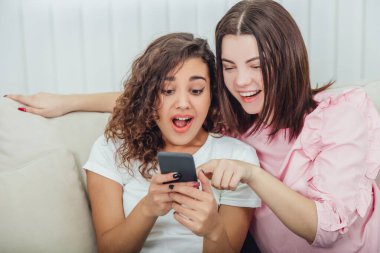 Evdeki kanepede oturan güzel genç kızlar telefondaki resimlere bakıp gülümsüyor, etkileşime geçiyorlar. Bir kız telefonu tutuyor, diğeri de ekranı işaret ediyor..