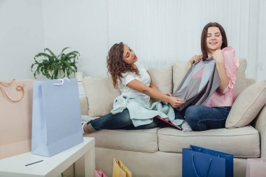 Genç kızlar dükkandan geri geldi. Kanepede oturuyorlar, çantalardan giysileri çıkarıp deniyorlar. Oda alışveriş paketleriyle dolu..