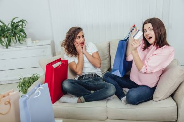 Kanepede oturan arkadaşlar. Bir kız gülümsüyor, elinde mavi bir çanta ve kredi kartı tutuyor, diğeri ise şaşkın bir ifadeyle ona bakıyor..