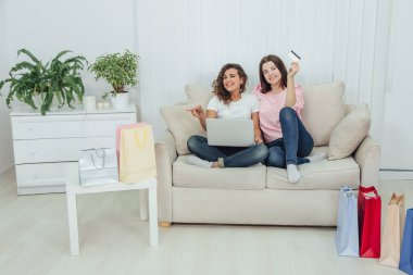 İnternetten alışveriş yapan iki mutlu bayan arkadaş. Bir kız kredi kartı tutuyor, diğeri de odadaki sayısız çöp torbasını işaret ediyor..