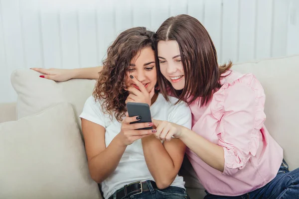 Beautiful young girls sitting on the sofa, at home, looking at the pictures in the phone, smiling, interacting. One girl is holding phone, another is pointing her finger at the screen. — Stock Photo, Image