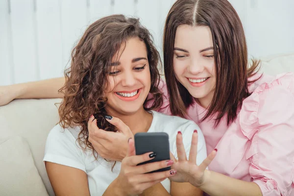Beautiful young girls sitting on the sofa, at home, looking at the pictures in the phone, smiling, interacting. One girl is holding phone, another is pointing her finger at the screen. — Stock Photo, Image