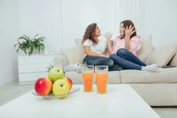 Montón de manzanas frescas y deliciosas y dos vasos de jugo en la mesa en la vanguardia. Dos chicas increíbles sentadas en el sofá sobre un fondo borroso, comunicándose emocionalmente en un ambiente hogareño . — Foto de Stock