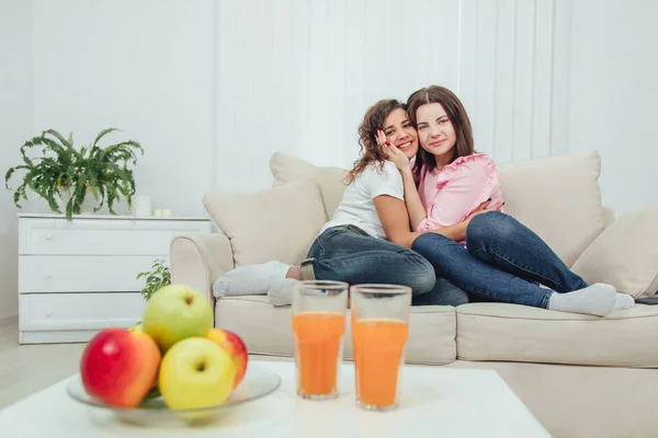 Dos chicas increíbles sentadas en el sofá, abrazándose, sonriendo encantadoras, mirando a la cámara. Manzanas borrosas y vasos de jugo en primera línea . — Foto de Stock