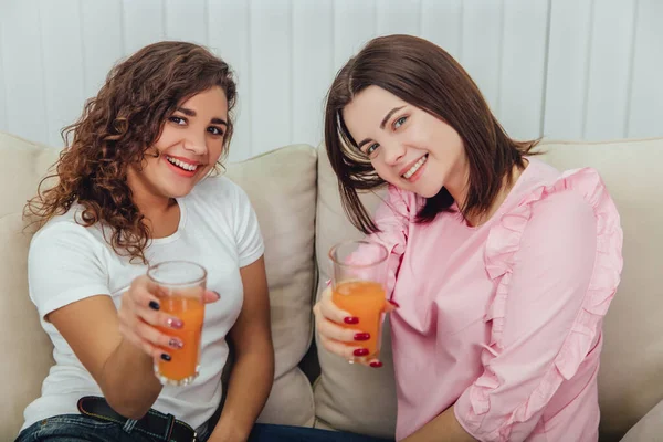 Afroamerikanische und kaukasische Freundinnen sitzen auf dem Sofa, trinken frischen Saft, posieren und lächeln in die Kamera. — Stockfoto
