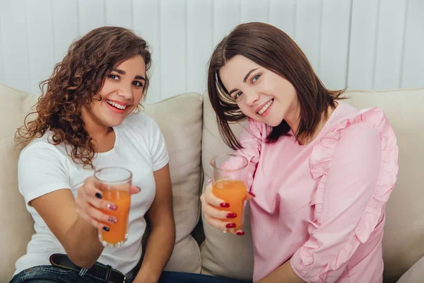 Afro-americanas y caucásicas amigas sentadas en el sofá, bebiendo jugo fresco, posando y sonriendo a la cámara . — Foto de Stock