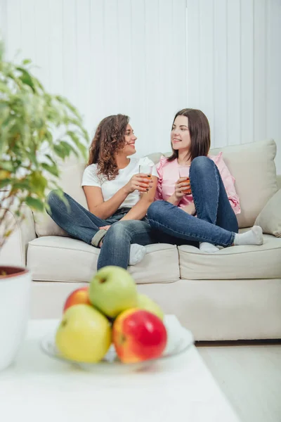 Placa borrosa de manzanas y maceta en primera línea. Dos chicas jóvenes lindas sentadas en el sofá con vasos de jugo en las manos, comunicándose, en el fondo — Foto de Stock
