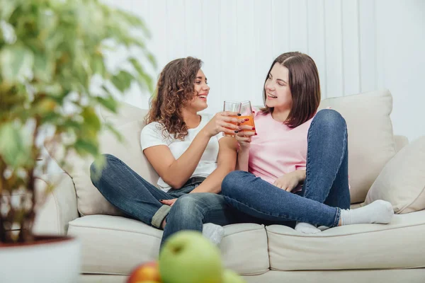 Placa borrosa de manzanas y maceta en primera línea. Dos chicas jóvenes lindas sentadas en el sofá tintineo vasos con jugo en el fondo . —  Fotos de Stock
