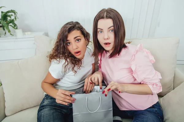 Two amazing friends sitting on the sofa, looking in one of the packets from the shop, surprised by something they have seen. — Stock Photo, Image