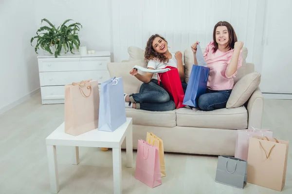 Extremely happy schopahollic girls sitting on the sofa, taking out clothes from colorful shop bags, smiling. — Stock Photo, Image