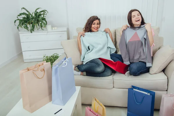Young girls came back from the shop. They are sitting on the sofa, taking clothes out from the bags and trying it on. Expressive satisfied face expressions. — Stock Photo, Image