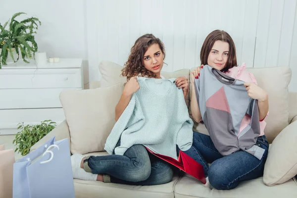 Молодые девушки вернулись из магазина. Они сидят на диване, вынимают одежду из мешков и примеряют ее. Выражающие удовлетворение выражения лица . — стоковое фото
