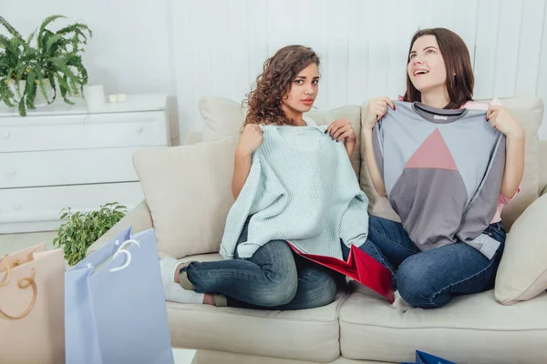 Young girls came back from the shop. They are sitting on the sofa, taking clothes out from the bags and trying it on. Expressive satisfied face expressions. — Stock Photo, Image