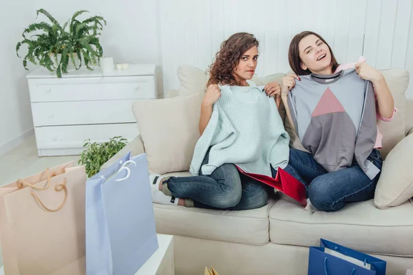 Young girls came back from the shop. They are sitting on the sofa, taking clothes out from the bags and trying it on. Expressive satisfied face expressions. — Stockfoto
