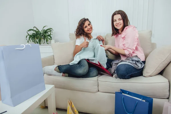 Young girls came back from the shop. They are sitting on the sofa, taking clothes out from the bags and looking at them, pointing thumb up. The room is full of shop packages. — Stock Photo, Image