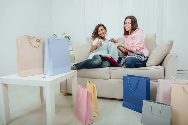 Young girls came back from the shop. They are sitting on the sofa, taking clothes out from the bags and looking at them, pointing thumb up. The room is full of shop packages. — Stock Photo, Image