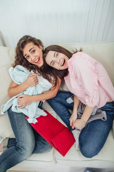 Duas garotas shopaholic sentadas no sofá, sorrindo, olhando para a câmera, desfrutando de roupas que compraram. Expressivas expressões faciais satisfeitas . — Fotografia de Stock