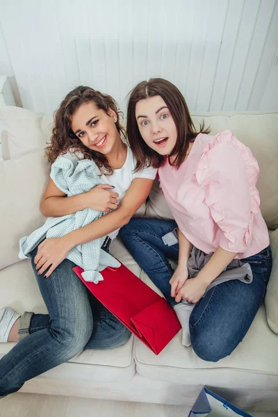 Dos chicas adictas a las compras sentadas en el sofá, sonriendo, mirando a la cámara, disfrutando de la ropa que han comprado. Expresiones faciales expresivas satisfechas . — Foto de Stock