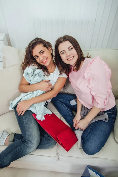 Zwei shopaholic Mädchen sitzen auf dem Sofa, lächeln, schauen in die Kamera und genießen Kleidung, die sie gekauft haben. Ausdruck zufriedener Mienen. — Stockfoto