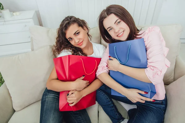 Duas garotas shopaholic sentado no sofá, sorrindo, olhando para a câmera, extremamente alegre, abraçando sacos de loja vermelho e azul. Expressivas expressões faciais satisfeitas . — Fotografia de Stock