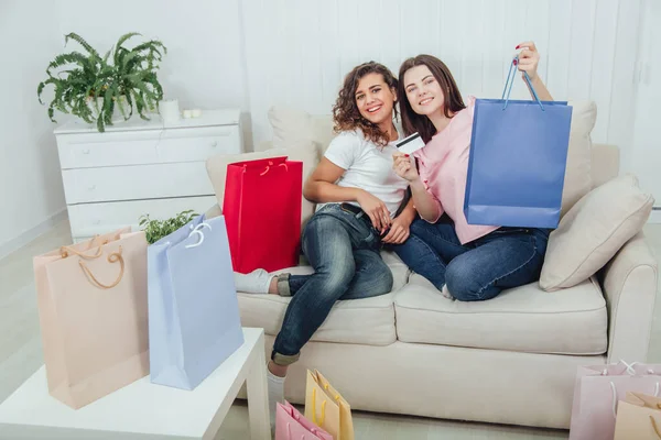 Cheerful girl friends sitting on the sofa, with bags from the shop all around the room and card in smiling brunettes hand. — Stock Photo, Image