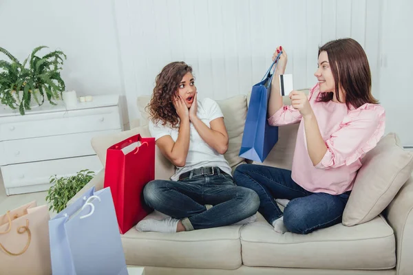 Amigos sentados en el sofá. Una chica está sonriendo, sosteniendo la compra en la bolsa azul de la tienda y la tarjeta de crédito, otra la está mirando con sorpresa, incluso confundida expresión de la cara . — Foto de Stock