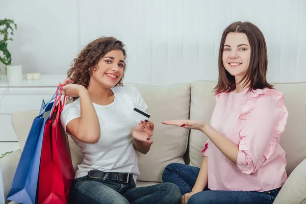 Friends sitting on the couch. One girl is smiling, holding purchases in blue and red shop bags and credit card, another is pointing at the card, looking at the camera. — Stock Photo, Image