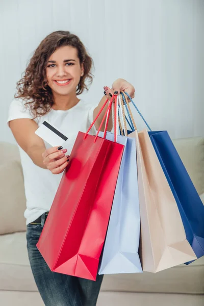 Niña rizada alegre borrosa con sonrisa brillante está extendiendo cuatro bolsas de tiendas de colores en una mano y tarjeta de crédito en otra a la cámara . — Foto de Stock