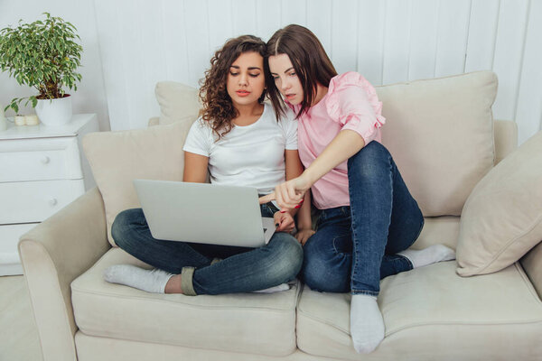 Two pretty young girls sitting on the sofa with laptop, surfing in the internet. Their legs crossed.