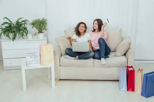 Due amiche felici che usano il computer portatile per fare shopping online. Una ragazza tiene in mano la carta di credito, un'altra punta il dito contro numerose borse spop in tutta la stanza. . — Foto Stock