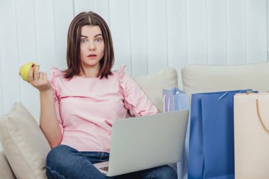 Kanepede loto pozisyonunda oturan sevimli bir kadın, elinde sarı elmayla internetten alışveriş yapmak için dizüstü bilgisayar kullanıyor, şaşırmış yüz ifadesiyle kameraya bakıyor..