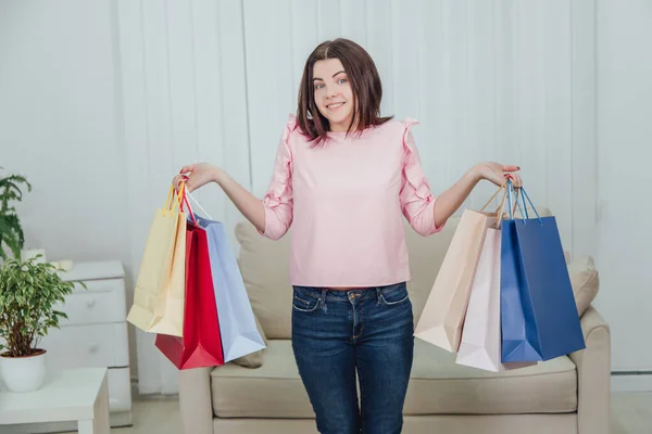 Young caucasian girl is standing with a lot of bags from shop in her hands, looking at the camera, smiling. — Stock Photo, Image