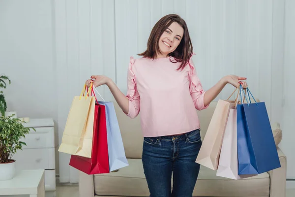 Young caucasian girl is standing with a lot of bags from shop in her hands, looking at the camera, smiling. — Stock Photo, Image