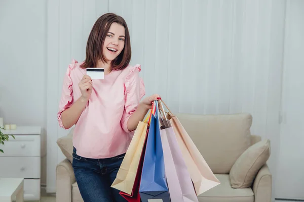 Young caucasian girl is standing with a lot of bags from shop and vredit card in her hands, looking at the camera, smiling. — Stock Photo, Image
