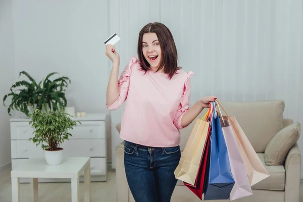 Young caucasian girl is standing with a lot of bags from shop and credit card in her hands, looking at the camera with excited face expression. — Stock Photo, Image