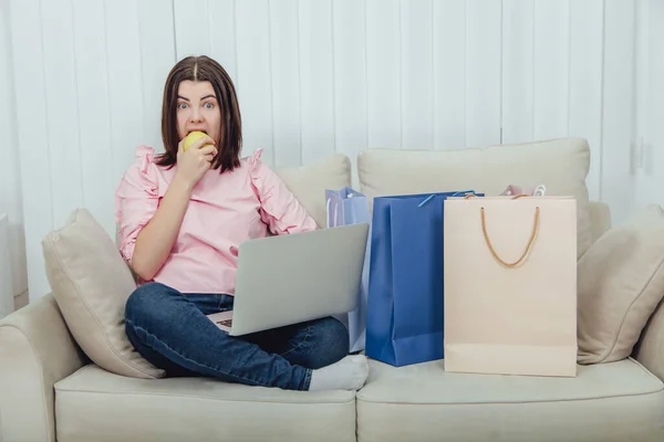 Niedliche Frau sitzt auf dem Sofa in lotos Position, Apfel essen, mit Laptop für den Online-Einkauf. Einkaufstüten in ihrer Nähe. — Stockfoto