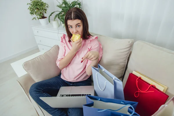 Glückliche Frau, die auf dem Sofa sitzt, Apfel isst, auf den Laptop zeigt, online einkauft. — Stockfoto