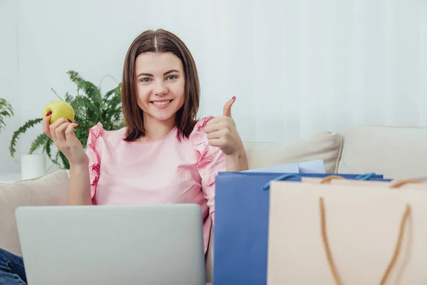 Glad kaukasisk dam håller äpple, använder laptop för att handla online, ger tummen upp, ler mot kameran. — Stockfoto