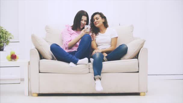 Jovens amigos bonitos sentados em um sofá na sala de estar, rindo alegremente de algo que estão assistindo no telefone . — Vídeo de Stock