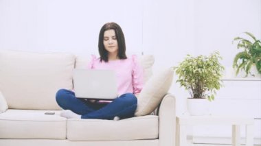 Kafkasyalı tatlı esmer kız loto pozisyonunda kanepede oturuyor, dizüstü bilgisayarla, internette sörf yapıyor, el sallıyor, kaşlarını kaldırıyor, kafası karışık görünüyor..