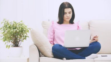 Kafkasyalı tatlı esmer kız loto pozisyonunda kanepede oturuyor, dizüstü bilgisayarla, internette sörf yapıyor, el sallıyor, kaşlarını kaldırıyor, kafası karışık görünüyor..