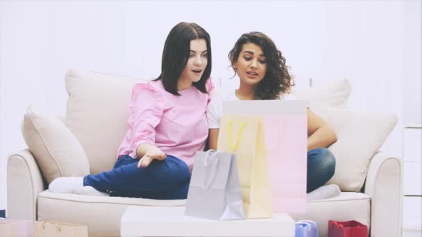 Dos amigas felices sentadas en el sofá, usando el portátil, eligiendo emocionalmente qué comprar en línea. Bolsas borrosas en la vanguardia . — Vídeo de stock