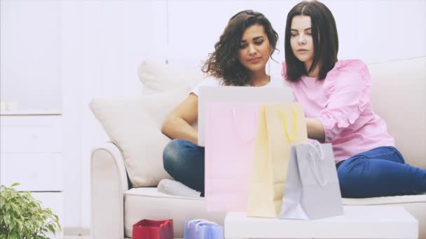 Dos amigas felices sentadas en el sofá, en casa, con portátil, eligiendo y discutiendo qué comprar en línea. Bolsas borrosas en la vanguardia . — Vídeos de Stock