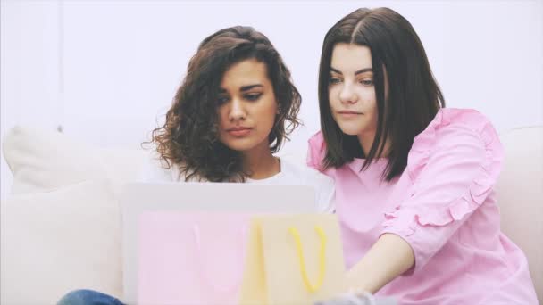 Dos amigas felices sentadas en el sofá, en casa, con portátil, eligiendo y discutiendo qué comprar en línea. Bolsas borrosas en la vanguardia . — Vídeo de stock