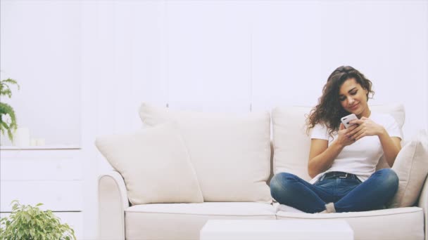 Menina afro-americana está sentada no sofá, com telefone nas mãos, mensagens. A amiga dela juntou-se a ela, trazendo muitos sacos da loja. Começam a olhar para as compras. . — Vídeo de Stock