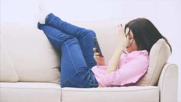 Nettes kaukasisches Mädchen liegt auf dem Sofa, legt die Beine auf die Rückseite des Sofas, benutzt Telefon, Nachrichten, lächelt, verdreht ihre Lippe. — Stockvideo