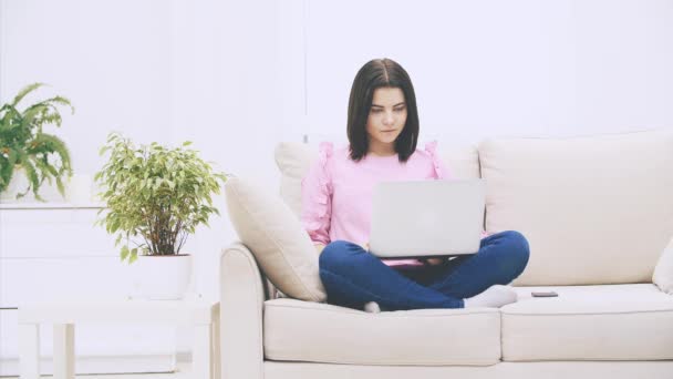 Schattig Kaukasisch brunette meisje zitten op de bank in lotos positie, met laptop, surfen op het internet. — Stockvideo